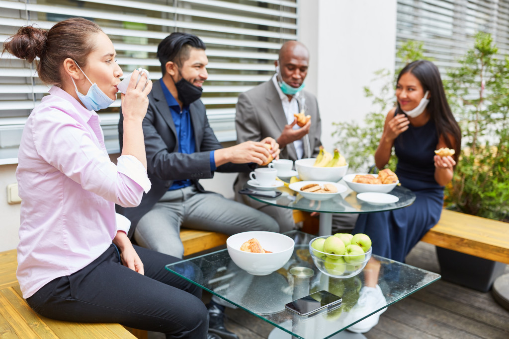 Young business people during a break eating together at the table with mouth-nose protection on the chin