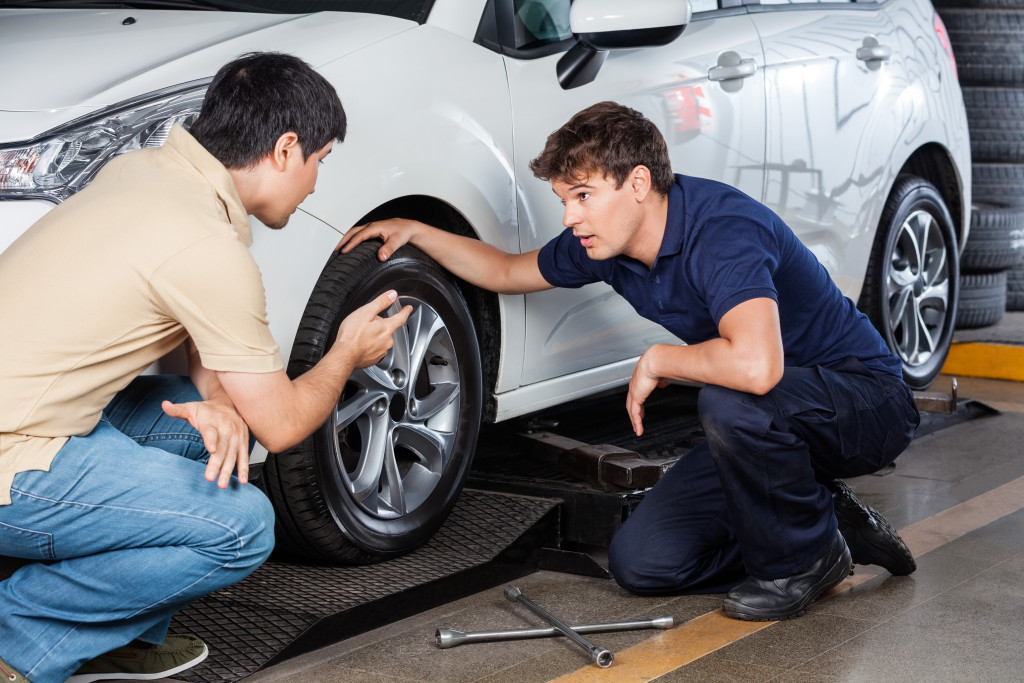 Auto repairman listens to customer's car issues while looking at the front tire of a car while at the auto shop